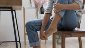 Woman at home holding her left foot due to pain caused by her Tailor's Bunion.